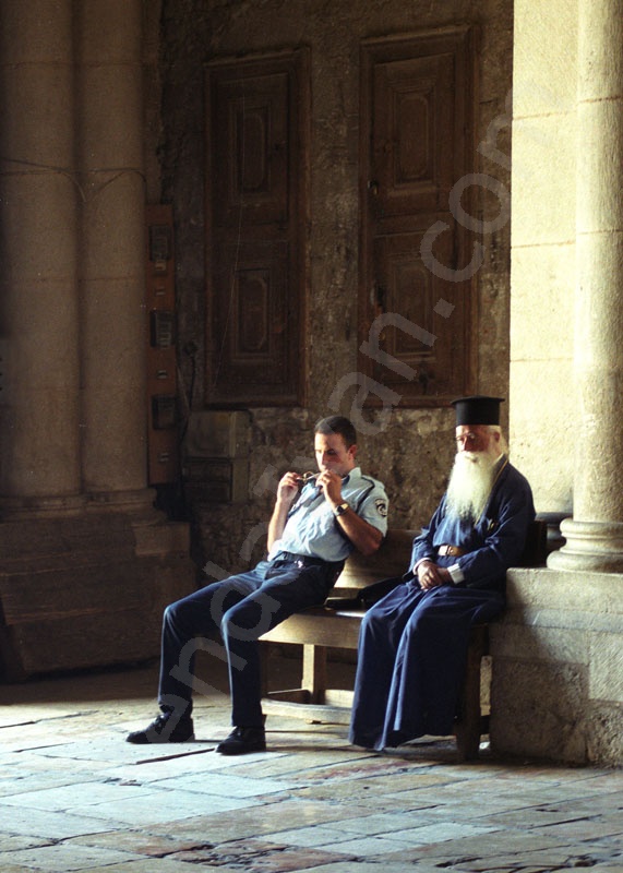  Szentsír templomban a bejárat melletti padon hosszú szakállas kék ruhás görög ortodox pap ül, izraeli rendőrrel.                 