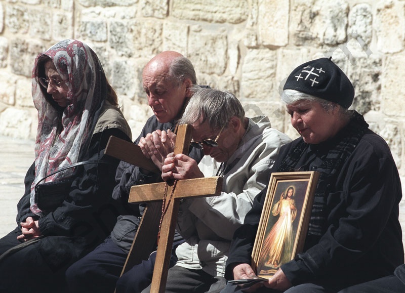 Örmény zarándokok csukott szemmel imádkoznak a Szentsír templom udvarán. Egyikük kezében kereszt van, a mellette ülő zarándok egy szentképet tart a kezében. 

             