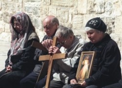 Örmény zarándokok csukott szemmel imádkoznak a Szentsír templom udvarán. Egyikük kezében kereszt van, a mellette ülő zarándok egy szentképet tart a kezében. 

             