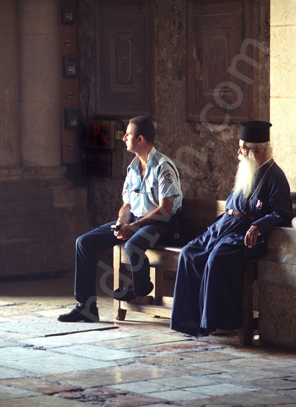     A bejárat melletti padon üll egy izraeli rendőr, és az idős szakállas görög ortodox pópa kék ruhában.                                      