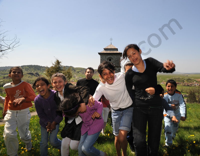       Vidám gyerekek játszanak a harangtorony előtt. Mögöttük a görög katolikus atya kedves mosollyal figyeli a gyerekeket. A völgyben Rakaca falu házai láthatók.                                      